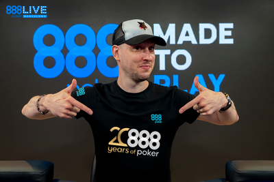 ian simspon -- a youngish white bald man wearing a baseball cap -- wears an 888poker shirt, pointing to the logo on the front with both hands. he stands in front of an 888poker banner.
