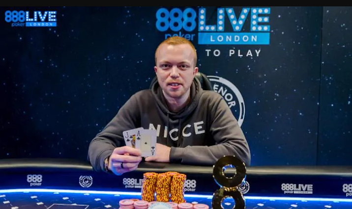 The winner of 888poker's London Live Series sits at a poker table holding up his winning hand: an ace and a jack of clubs. He has a pile of chips in front of him along with a trophy shaped like a giant 8. More than 600 players competed in the Main Event.