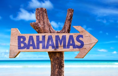 a wooden sign is attached to a wooden pole.  The sign is arrow-shaped and says "BAHAMAS".  behind, you can see sunny blue skies, sand and bright blue waters.