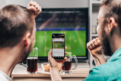 Two freinds are watching sports on TV & cheering over a play. One of them holds a cell phone in one hand with the BetMGM Sportsbook app live betting feature on the sceen