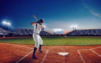 A professional baseball player is seen up at bat from behind, he holds the bat firmly behind him ready to swing, in the dark, you can see the stadium full of fans anxiously waiting.