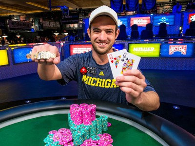 WSOP Main Event winner and WSOP MI brand ambassador Joe Cada is seen smiling at a poker table, holding up a pair of queens and showing off his WSOP gold bracelet. A pile of pink and teal poker chips are on the table in front of him.