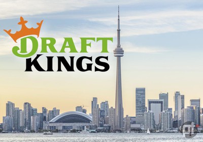Toronto skyline is seen against blue sky. CN Tower and skyscrapers rise up from the shore of Lake Ontario. On the upper left hand corner is the DraftKings Logo.