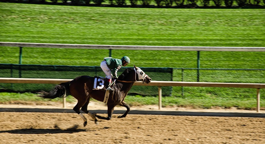 A racehorse is seen racing down the track in Kentucky, wearing the number 13. The jockey riding him wears all green.  A new Kentucky bill would provide for online and retail sportsbook in conjunction with the state's 7 horserace tracks.