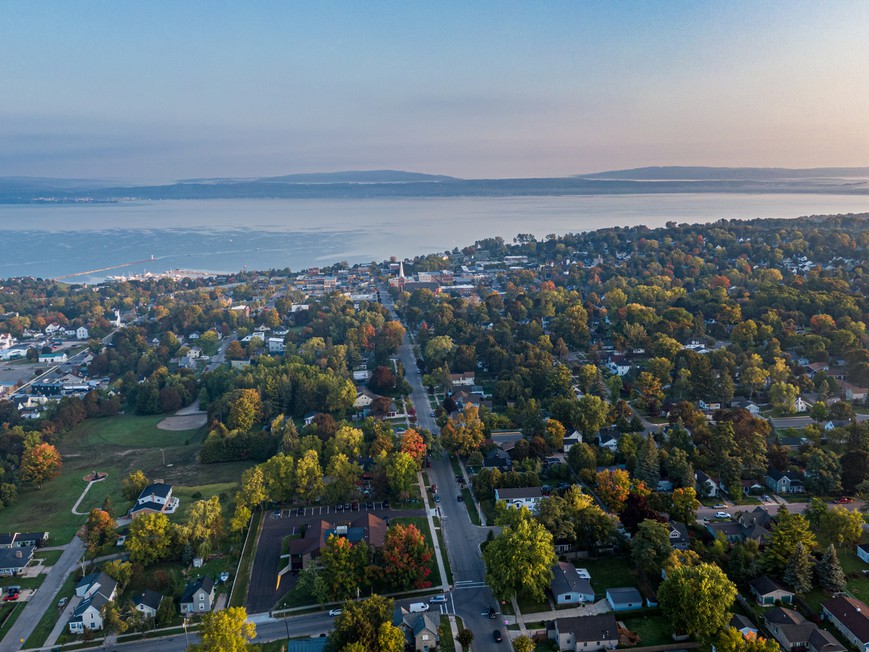 Lake Michigan is seen from above, Rows of trees and houses in the foreground lead to a gorgeous expanse of water with a pink and purple haze on the horizon. 