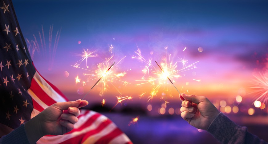 an american flag is seen on the left. next to it, two hands holding sparklers reaching towards each other in the middle of the frame in front of a dark nighttime background.