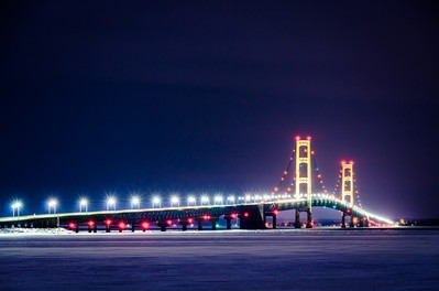 Mackinac Bridge is seen at night, illuminated and reflected in the dark water below as it stretches above.  Mackinac Bridge is a suspension bridge that crosses the Mackinac Strait and connects the upper and lower peninsulas of the US state of Michigan.