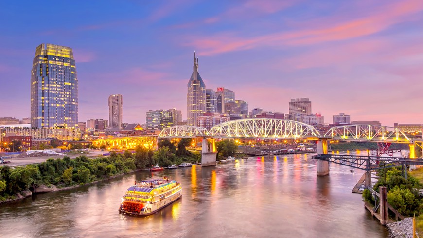 Nashville, Tennessee downtown skyline at twilight. As the sun sets pink and blue, you see it reflected in the river as a boat drives down. The bridge connects the land full of trees and buldings.