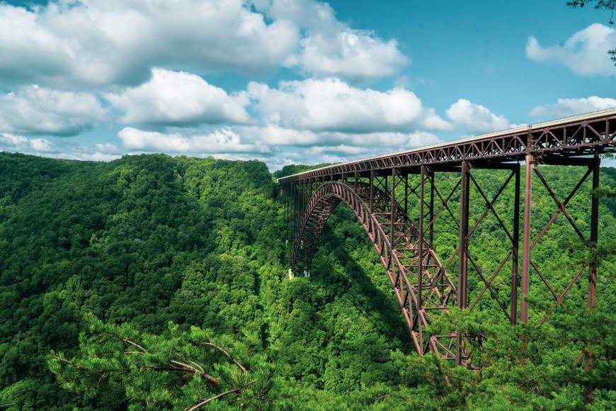  beautiful elevated train track is seen going into the lush mountains of West Virginia below a blue sky with puffy white clouds.