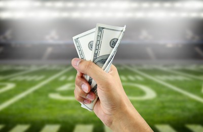 a hand holding two crisp $100 bills in front of an American Football field. Score Big with NFL 2023/24: Best New US Sportsbook Bonuses!