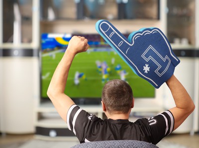 A football fan wearing a jersey and a foam finger cheers while watching an NFL game on the tv. Turn $5 into $390 with NFL Preseason Picks & Win Big this Weekend