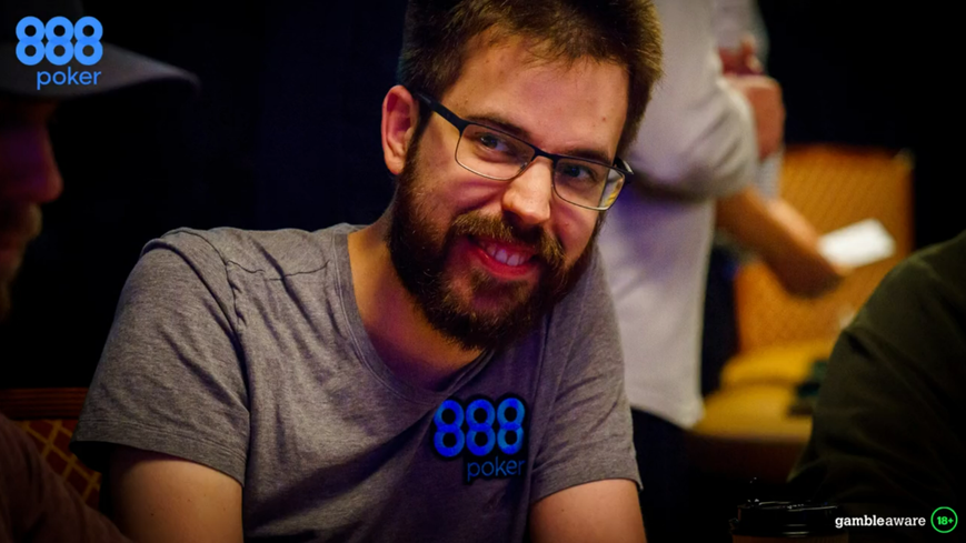 Dominik Nitsche is seen wearing a heather grey 888poker t-shirt seated at a poker table at a tournament. He has slightly poofy hair, black thin framed glasses, and a beard. Dominik Nitsche exits the stage after seven years with 888poker, but the operator 