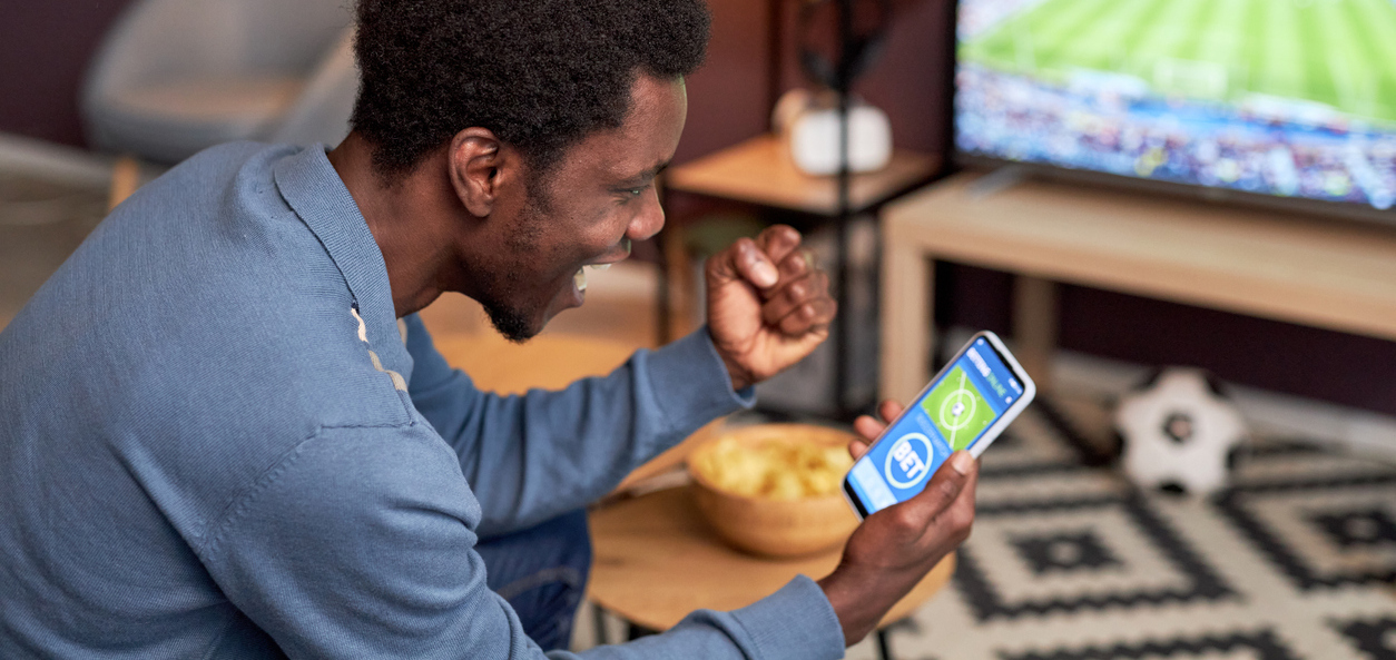 Side view portrait of black man holding smartphone with online sport bets app and cheering for football team.