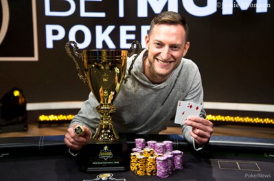a smiling white man is sitting at a poker table.  he brandishes an ace and a four of diamonds.  next to him is a large gold trophy and a stack of poker chips.