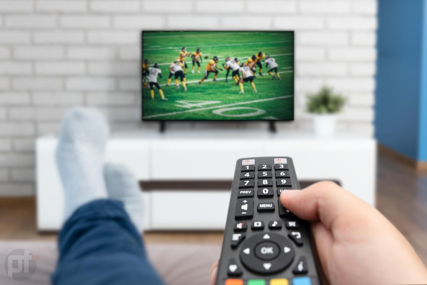 a man holding a remote as he watches football on the TV. Where to Watch Sports Thanksgiving Weekend 2023